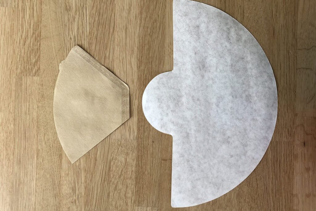 Ein trapezförmiger brauner Kaffeefilter neben einem halbrunden Chemex-Filter auf Holz