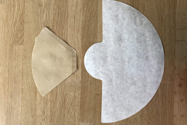 Ein trapezförmiger brauner Kaffeefilter neben einem halbrunden Chemex-Filter auf Holz