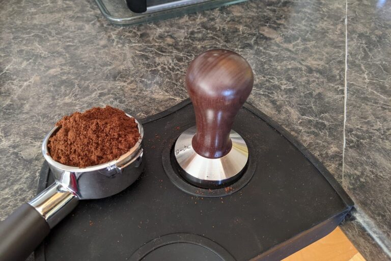 Siebträger mit frisch gemahlenem Kaffee neben einem Tamper auf einer Tampermatte